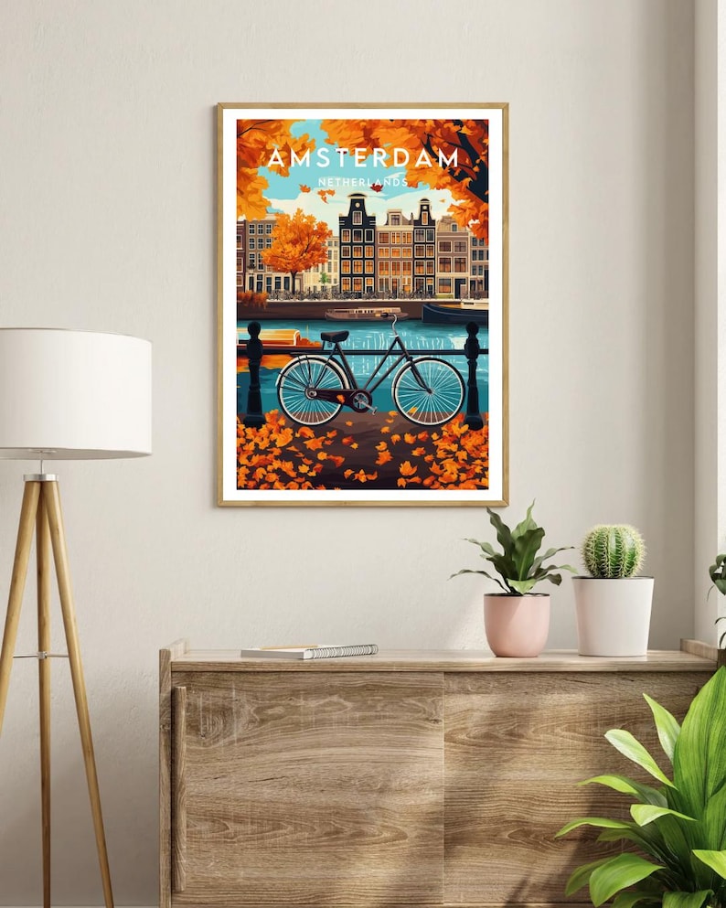 May include: A vintage travel poster featuring a bicycle parked in front of a canal in Amsterdam, Netherlands. The poster has a blue, green, and orange color scheme with fall foliage in the foreground.