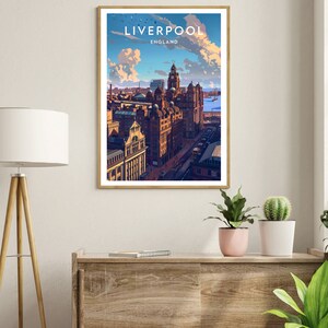 May include: A framed art print featuring a stylized illustration of Liverpool, England. The artwork showcases a cityscape with prominent buildings under a blue sky with clouds. The print is in a wooden frame and displayed on a wooden cabinet.