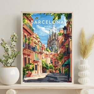 May include: A framed illustration of a street scene in Barcelona, Spain. The illustration features a narrow street lined with colorful buildings, with the iconic Sagrada Familia cathedral visible in the background. The sky is blue and sunny, and there are trees and plants growing along the street.