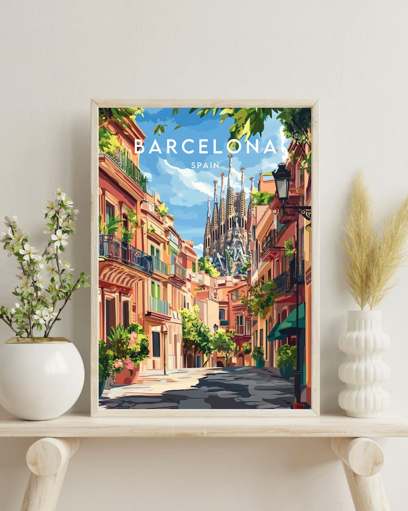 May include: A framed illustration of a street scene in Barcelona, Spain. The illustration features a narrow street lined with colorful buildings, with the iconic Sagrada Familia cathedral visible in the background. The sky is blue and sunny, and there are trees and plants growing along the street.