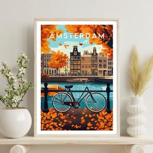 May include: A vintage travel poster featuring a bicycle parked by a canal in Amsterdam, Netherlands. The poster has a blue and green color scheme with orange and yellow fall leaves scattered on the ground. The text on the poster reads "AMSTERDAM, NETHERLANDS".