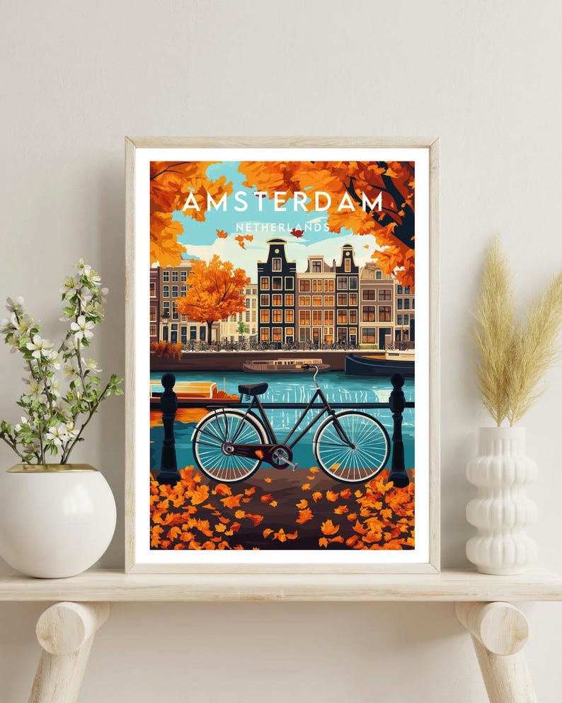 May include: A vintage travel poster featuring a bicycle parked by a canal in Amsterdam, Netherlands. The poster has a blue and green color scheme with orange and yellow fall leaves scattered on the ground. The text on the poster reads "AMSTERDAM, NETHERLANDS".