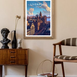May include: A framed art print featuring a stylized illustration of Liverpool, England, with the city's skyline under a blue sky with fluffy clouds. The print is in a wooden frame and hangs on a beige wall. The print has the word "LIVERPOOL" at the top.