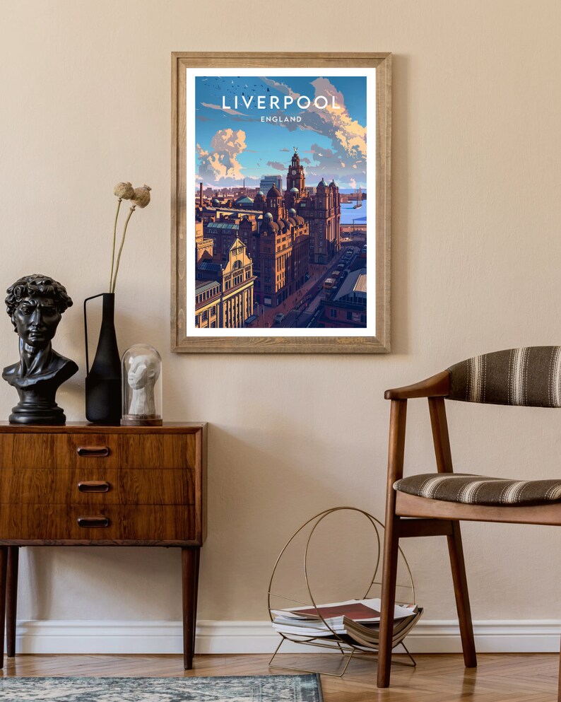 May include: A framed art print featuring a stylized illustration of Liverpool, England, with the city's skyline under a blue sky with fluffy clouds. The print is in a wooden frame and hangs on a beige wall. The print has the word "LIVERPOOL" at the top.