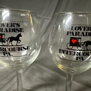 May include: Two clear wine glasses with a stem. Each glass has the words "LOVER'S PARADISE" above a graphic of a horse-drawn carriage with a red heart and the words "INTERCOURSE PA." in black.