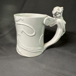 May include: A light-colored ceramic mug featuring a sculpted handle shaped like a winged figure. The body of the mug is decorated with embossed, flowing organic lines and plant-like motifs that wrap around the exterior surface of the vessel.