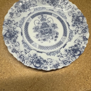 May include: White ceramic plate with a scalloped edge, adorned with a blue floral pattern. The design features roses and other flowers, with a central floral bouquet and a patterned border. A decorative piece for serving.