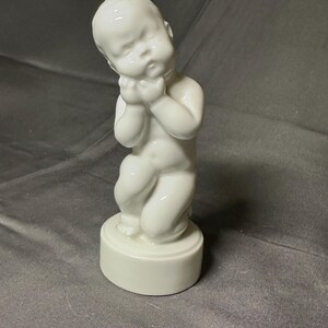 May include: A white ceramic figurine of a baby kneeling with its head resting on its hands. The baby is on a round base. The figurine has a glossy finish and is set against a gray background.