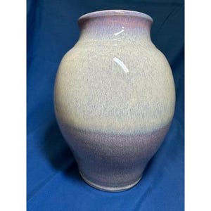 May include: A handcrafted ceramic vase with a wide, rounded body and a slightly flared rim. The vase features a two-tone color scheme, with a lavender-purple base that fades into a speckled, off-white and lavender upper section. The vase has a glossy finish.