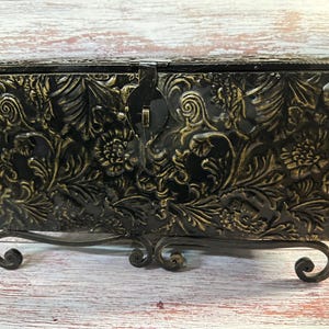 May include: Black decorative chest with a floral embossed design and a metal latch. The chest sits on a decorative metal stand with scrolled feet. The chest is approximately 12 inches long, 8 inches wide, and 6 inches tall.