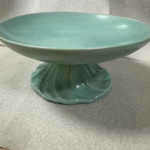 May include: A turquoise ceramic pedestal bowl. The bowl has a wide, shallow dish supported by a decorative, swirled base. The color is a muted, seafoam green. The bowl is likely vintage and suitable for displaying fruit or other decorative items.