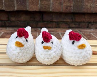 Könnte beinhalten: Drei kleine, runde, gehäkelte Hühner aus weichem, strukturiertem Garn. Jedes Huhn hat einen leuchtend roten, gehäkelten Kamm auf dem Kopf, einen kleinen orangefarbenen Schnabel und zwei schwarze Perlenaugen. Die Hühner stehen in einer Reihe.