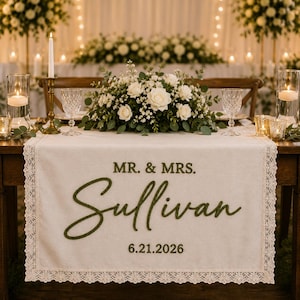 May include: A cream-colored fabric table runner with lace trim featuring the embroidered text MR. & MRS. Sullivan and the date 6.21.2026 in dark green thread. The runner is displayed on a wooden table with a floral centerpiece and candles.