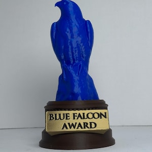 Könnte beinhalten: Eine blaue Falkenfigur auf einem dunkelbraunen, runden Sockel. Der Sockel ist mit einem rechteckigen, goldfarbenen Schild versehen, auf dem der schwarze Text "BLUE FALCON AWARD" zentriert ist. Der Falke ist in einer aufrechten Position dargestellt.