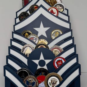Puede incluir: Un expositor de madera azul marino y blanco con forma de galones militares, que contiene varias monedas de desafío. El expositor presenta una gran estrella blanca y contiene monedas de diferentes colores y diseños, incluyendo temas militares y patrióticos.