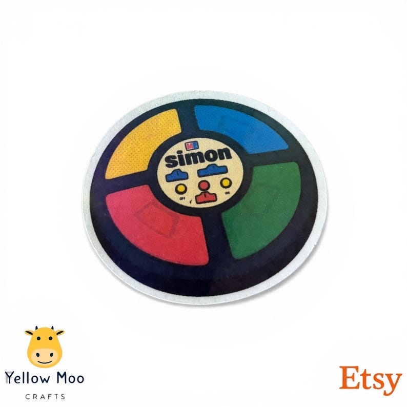 Pu&ograve; includere: Un pulsante circolare del gioco Simon con un bordo nero e un bordo argentato. Il pulsante presenta sezioni gialle, blu, verdi e rosse. La parola "Simon" &egrave; stampata in bianco, con una piccola grafica di un volto.