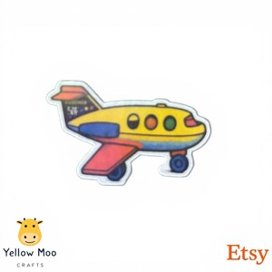 Pu&ograve; includere: Un bottone a forma di aereo colorato con corpo giallo, ali rosse e blu e ruote blu. Il bottone ha un design a cartoni animati con dettagli arancioni, verdi e marroni. La parola "Passenger" &egrave; visibile sulla coda.