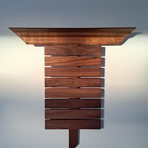 Op de afbeelding: Een wandplank van hout met een brede, horizontale bovenplank met afgeschuinde randen. Onder de plank zijn acht horizontale houten latten in een verticale stapel geplaatst, bevestigd aan een centrale verticale steun van donkerbruin hout.