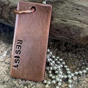 May include: A copper-colored rectangular pendant necklace with the word "RESIST" stamped on it. The pendant hangs from a silver ball chain. The pendant has a small loop at the top for the chain to pass through.