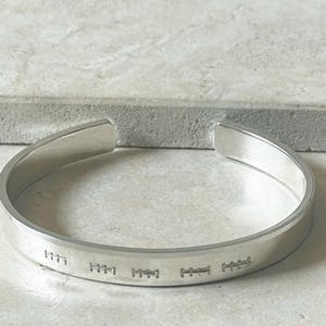 Puede incluir: Una pulsera de plata con marcas grabadas. La pulsera abierta tiene un acabado liso y pulido y un diseño simple y moderno. Los detalles grabados están espaciados uniformemente alrededor de la banda.
