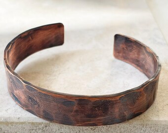 Puede incluir: Un brazalete de cobre martillado. El brazalete abierto tiene una superficie texturizada con tonos marrón oscuro y marrón rojizo. El brazalete mide aproximadamente 6,35 cm de ancho.