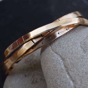 May include: Three gold bracelets with a hammered finish. The bracelets are stacked on top of each other and have an arrow design engraved on them.