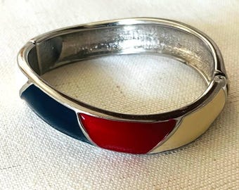 May include: A silver-coloured hinged bracelet with a tri-colour design. The bracelet features sections of navy blue, red, and cream-coloured enamel. The bracelet is shown on a white, textured surface.