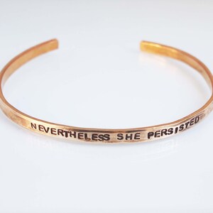 Puede incluir: Un brazalete dorado con la inscripción "Nevertheless she persisted".