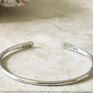 Puede incluir: Una pulsera de plata con textura martillada. La pulsera es abierta, con las iniciales "A & L" y un símbolo de corazón estampados en un extremo, y la fecha "6.6.09" en el otro. Joyería.