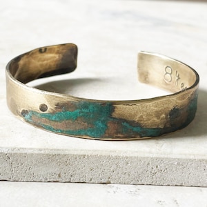 May include: A gold-toned metal cuff bracelet with a hammered texture and a prominent oxidized teal and brown patina across the front surface. The interior of the bracelet features the stamped text 8 years, visible on the right side of the opening.