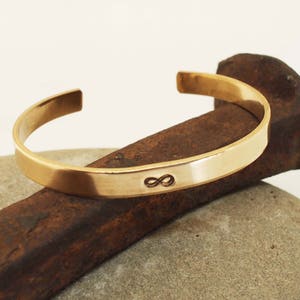 Puede incluir: Un brazalete de metal dorado con un símbolo de infinito grabado en la parte delantera.