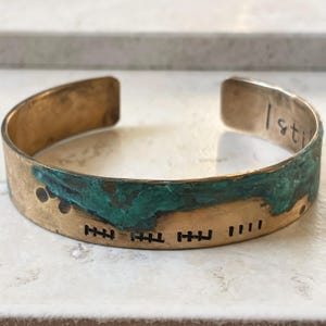 Puede incluir: Una pulsera de latón con un acabado envejecido y una pátina turquesa. La pulsera presenta marcas grabadas y la palabra "I still" en el interior. La pulsera es abierta y ajustable.