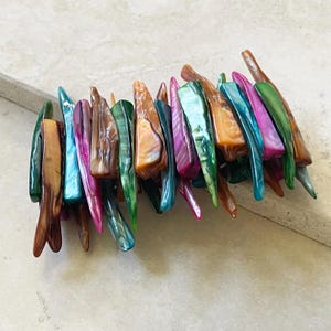 Puede incluir: Una colorida pulsera compuesta por cuentas alargadas de forma irregular. Las cuentas muestran una gama de colores, incluyendo marrón, verde, azul, rosa y naranja, con una calidad brillante e iridiscente. La pulsera está diseñada para ser usada alrededor de la muñeca.