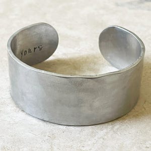 May include: A wide, silver-colored cuff bracelet. The bracelet has a brushed metal finish and is open at the ends. The word "Yours" is inscribed on the inside of the bracelet.