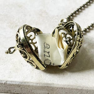 May include: A heart-shaped, antique gold locket necklace with an openwork design. The locket is open, revealing a small, folded piece of paper with handwritten text. The necklace chain is a matching antique gold color.