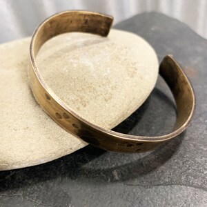 May include: A hammered brass cuff bracelet with a smooth, polished finish. The bracelet is open ended and has a slightly curved shape.