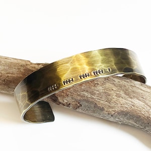 May include: A brass cuff bracelet with a hammered finish. The bracelet has a simple inscription that reads "HI MY THY THE I".