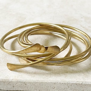 Peut inclure: Un ensemble de bracelets de couleur or. Les bracelets sont faits de métal fin et incurvé et sont entrelacés. L'ensemble comprend un fermoir avec un motif décoratif en forme de feuille. Les bracelets sont présentés sur une surface de couleur claire.