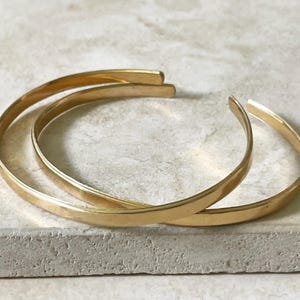 Puede incluir: Tres pulseras de puño doradas. Las pulseras son bandas curvas simples con un acabado liso y pulido. Están apiladas una encima de la otra, creando un efecto en capas. Las pulseras se exhiben sobre una superficie clara y texturizada.