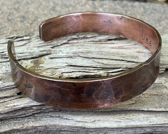 Puede incluir: Una pulsera de puño de cobre con textura martillada. La palabra "One" está estampada a mano en el exterior. La pulsera mide aproximadamente 2 cm de ancho.