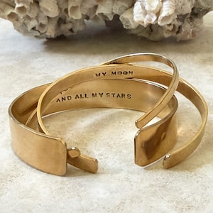 Peut inclure: Deux bracelets joncs en métal doré à extrémités ouvertes. La surface intérieure de l'un des bracelets est gravée du texte MY MOON, tandis que l'autre bracelet porte le texte gravé AND ALL MY STARS dans une police majuscule sans empattement.