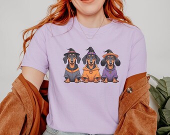 May include: A light purple t-shirt featuring three dachshunds dressed in Halloween costumes. Each dog wears a witch hat and a different colored hoodie: gray, orange, and purple. The dogs are sitting and facing forward.