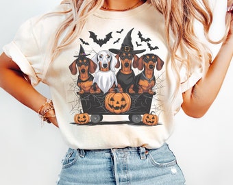 May include: Ivory-colored t-shirt featuring a Halloween-themed graphic. The design showcases four dachshunds in costumes, riding in a cart with a carved pumpkin. Bats and spiderwebs complete the spooky scene. The word "IVORY" is printed at the top.