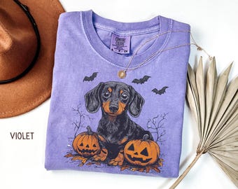 May include: Violet-colored t-shirt featuring a dachshund dog illustration with two carved pumpkins and bats. The shirt has a crew neck and short sleeves. A gold necklace is draped across the shirt. The word "VIOLET" is printed in the lower left corner.