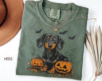 May include: Moss green Comfort Colors t-shirt featuring a dachshund dog illustration with two jack-o'-lanterns and bats. The shirt has a crew neck and short sleeves. The shirt is a size small.