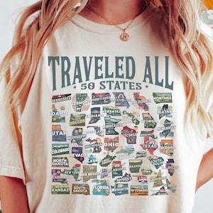 May include: A cream-coloured t-shirt with the text "Traveled All *50 States*" printed on the front. The shirt features a colourful graphic of the United States with each state represented by a small, illustrated patch.