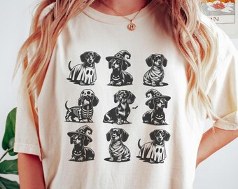 May include: Cream-colored t-shirt featuring a grid of nine dachshund illustrations in Halloween costumes. The dachshunds are depicted as a ghost, witch, mummy, and skeleton. The shirt is a comfortable, casual style.