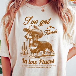May include: A white t-shirt featuring a brown dachshund wearing a cowboy hat and the text "I've got Friends in low Places". The text below the image reads "All I need is my dachshund, the shortest littlest cutest living creature in the world #friendsinlowplaces".