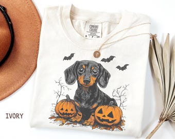 May include: Ivory-colored t-shirt featuring a black and brown dachshund dog illustration with two carved pumpkins and bats. The shirt has a Comfort Colors tag and a gold necklace. The word "IVORY" is printed on the left side.