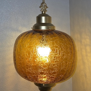 May include: A vintage amber-colored pendant light fixture. The globe-shaped glass shade has a textured, crackled appearance. The fixture includes a brass-toned metal cap, chain, and decorative accents. The light is turned on, illuminating the interior.